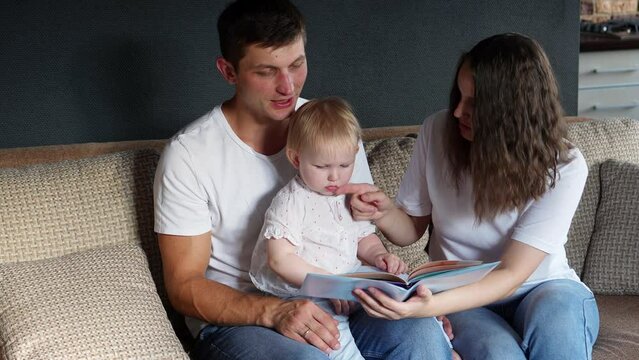 Caring parents read a book to their child and show pictures while sitting on couch at home. Young loving parents spend time with their adorable baby, learning and communicating. Happy family concept. 