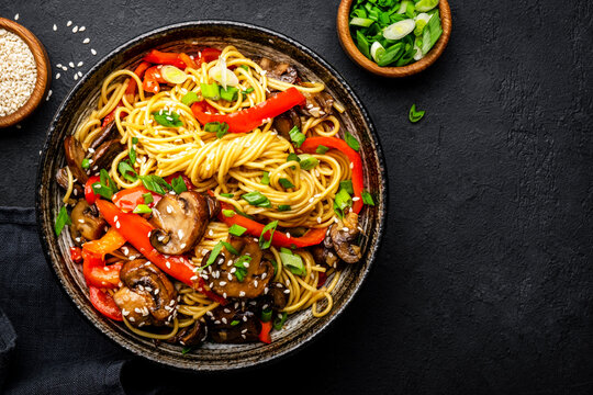 Vegan stir fry noodles with red paprika, champignons, green onion and sesame seeds with ginger, garlic and soy sauce.. Black table background, top view