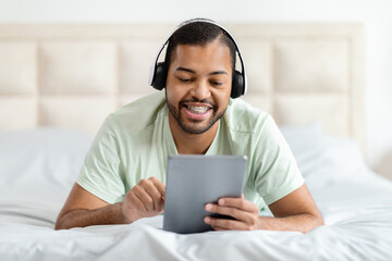 Happy black man using digital tablet and headphones in bed