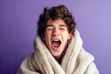 Fotobehang A young man with his mouth wide open, wrapped in a blanket, appears sleepy on a lilac background. © Joaquin Corbalan