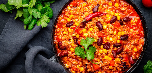 Chili con carne, mexican dish with minced beef, red beans, paprika, corn, cilantro and red peppers in spicy tomato sauce, tex-mex cuisine, black table background, top view banner