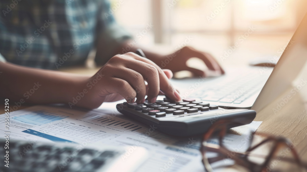 Poster close-up view of a person's hands working on a financial report with a calculator and a laptop in th