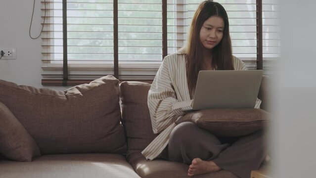 Asian woman sitting on sofa working from home using laptop
