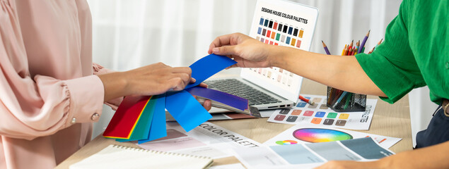 Cropped imaged of professional interior designers choose appropriate color from color palette with house model and architectural equipment scatter around. Closeup. Focus on hand. Variegated.