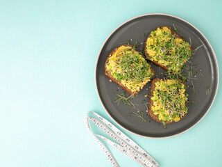 A snack of black bread with avocado and arugula sprouts, a micrograin on a gray plate and a measuring tape on a blue one. The concept of proper nutrition, low-calorie snacks and weight loss