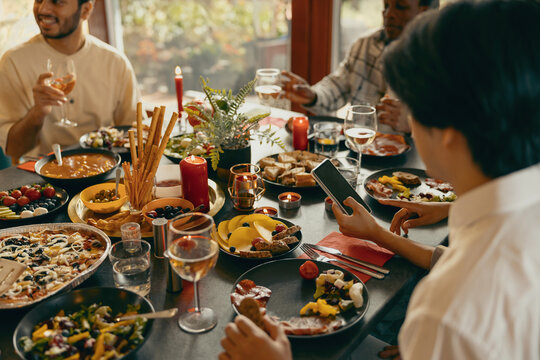 Close up of friends sitting at festive dinner table at home, drinking wine and looking at phone