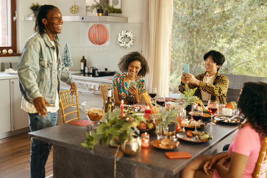 Smiling men and women sitting at holiday dinner table at home, eating and use mobile phone
