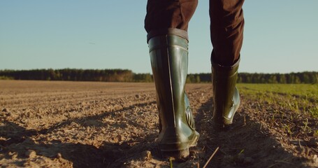 Low angle: man walking in rubber boots in a farmer's field, the blue sky above the horizon. Man walking through an agricultural field. Farmer walks through a plowed field in early spring. - 755885075