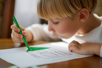 Small kid drawing a house on a table. Young caucasian boy sketching with markers at home on a sunny day. Blonde child coloring a sketch of a house with markers as his homework assignment.
