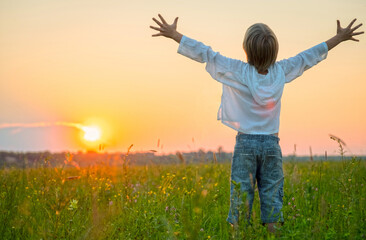 Happy boy with raised hands looks at the sunset  in summer. Boy in a field with his hands raised while sunrise. Freedom and relaxation concept. Blonde kid looking to the sun at the evening. - 755886666