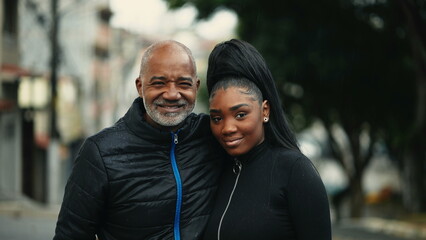 Middle-aged father and teen daughter posing together for camera standing outside in urban environment during drizzle rain. South American hispanic people of African descent