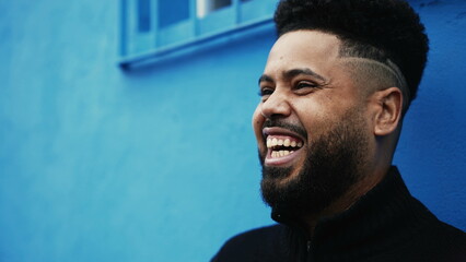 One happy young black Brazilian man laughing and smiling in front of an urban blue wall, close-up face of a South American person conversing with friends off-camera - obrazy, fototapety, plakaty