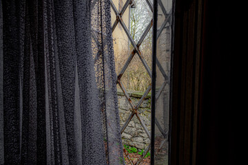 Aussicht auf eine Mauer, Gebäude und Bäume durch ein vergittertes Fenster mit weißem altertümlichen Vorhang