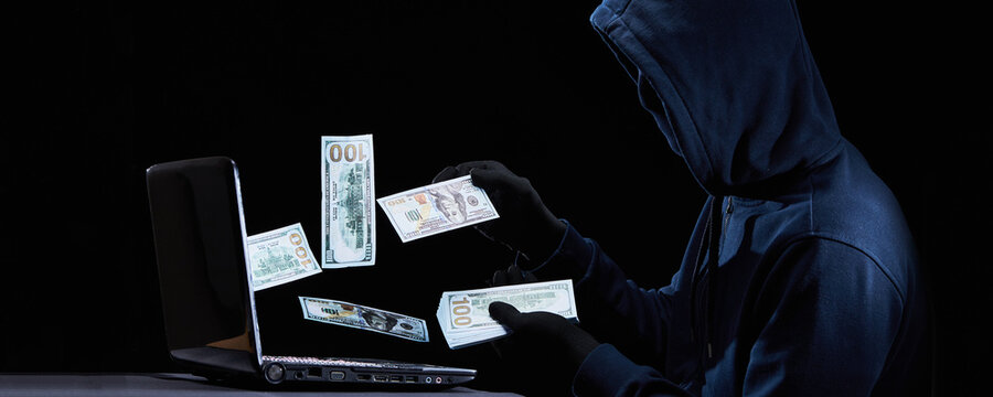 A hacker in a hoodie and a black mask steals money using a laptop and the Internet. The concept of Internet theft, cybercrime, attacks on digital data.