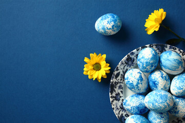 Plate with blue and white Easter eggs and yellow flowers on dark blue table. Top view.