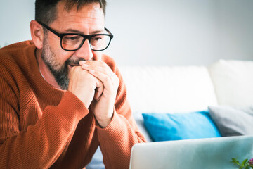 Focused young Caucasian man in glasses sit on sofa at home work online on laptop gadget....