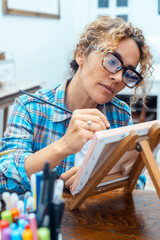 One woman alone at home enjoying indoor leisure activity painting a canvas. Mature artist enjoy creative hobby. Picture of serious concentrated Caucasian female artist sitting at desk. Wellbeing relax