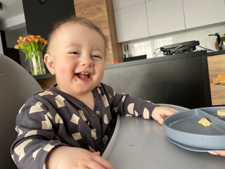 Portrait of a baby boy who looks into the camera and sincerely laughs while sitting at the feeding...