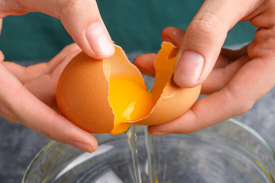 Housewife hands cracking fresh egg, yolk and white dropping in a bowl. A woman's hand cracking an egg with knife into a clear glass bowl in the gray kitchen table, the process of cooking, close up
