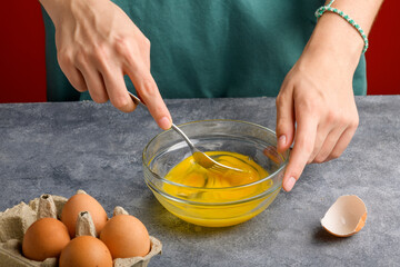 Female hands beat eggs with fork in a glass bowl on a gray kitchen table. Cooking scrambled fresh eggs for breakfast, the process of omelette cooking, close up