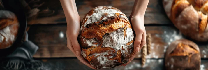 Keuken spatwand met foto Hands holding freshly baked bread above wooden table, showcasing homemade artisan sourdough with flour-dusted crust in natural light. Banner. Copy space © AI_images