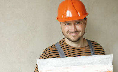 Smiling handyman repairman worker in overalls and protective orange helmet holds big putty knife...