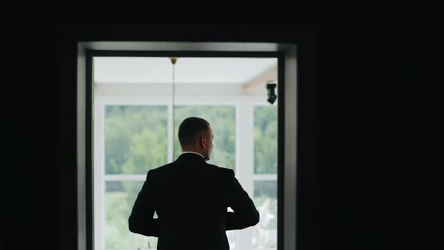 The silhouette of a man standing at the exit of the room and getting dressed. He adjusts his jacket on himself. Statuesque silhouette of a businessman