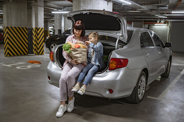 Clad in casual clothing, Caucasian mother and little daughter sitting in trunk of their vehicle,...