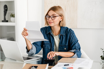 Young caucasian woman paying domestic bills, making calculations at home desk. Female holding...