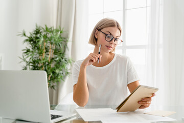 Young caucasian woman doing homework, taking notes using laptop at home. Female freelancer writing new ideas, creating startup, working remotely