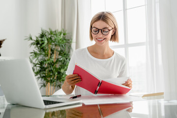 Happy young caucasian woman taking notes, writing and doing e-learning using laptop at home. Female student freelancer watching seminars webinars
