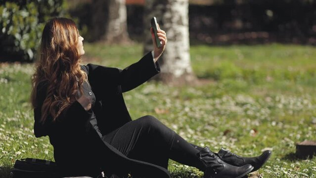 A beautiful girl, a student, takes a selfie on her phone in a park in a clearing with flowers and daisies. Spring mood, warmth, sunbathing, joy, beauty, bloom, social networks, technology