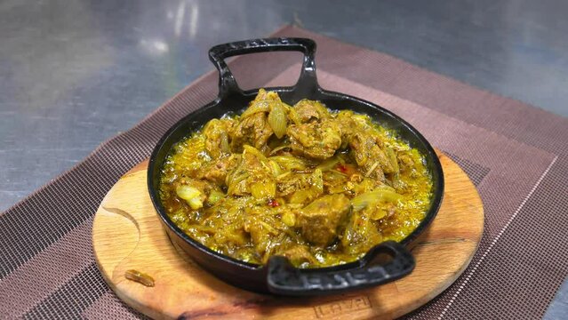 Chef in black glove in kitchen prepares Georgian dish Ostri, in cast iron pan on wooden board on table in restaurant . Adds meat in sauce to hot scoop.