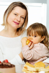 Obraz na płótnie Canvas Woman giving apple to a child for snack, sitting beside table full of sweet food and unhealthy pastry with chocolate. Concept of parents choice wtah to give her child to eat, healthy lifestyle