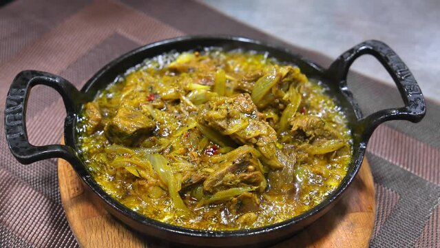 Chef in black glove in kitchen prepares Georgian dish Ostri, in cast iron pan on wooden board on table in restaurant . Adds herbs to meat in sauce to hot scocolike.
