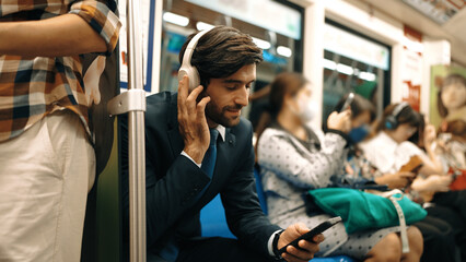Smiling handsome male investor listening relaxing music while sitting on train. Professional business man enjoy listening music while using public transport to workplace. Blurred background. Exultant.