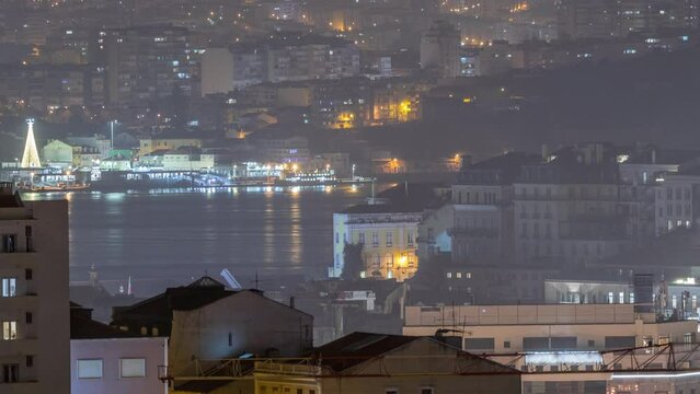 Aerial view of Lisboa downtown night timelapse. Panoramic of Baixa, Rossio and Chiado rooftops from above. Illuminated buildings. Almada on the other side of Tejo river. Portugal