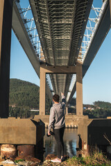 Young man with a camera in grey hoodie points across Rande Bridge from below. Pillars and highway stretching over Vigo Estuary, create perspective as the tranquil sea mirrors the scene on sunny day.
