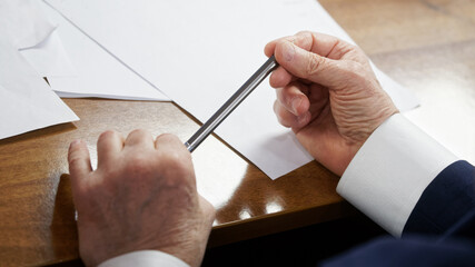 Elderly man sits at a table with white sheets of paper and holds a metal pen in his wrinkled hands....