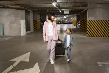 Young woman with child holding traveling suitcase as searching for car before embarking on trip. Caucasian mother and little daughter dressed in casual clothes walking through underground parking lot.