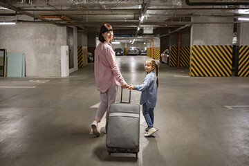 Fototapeta na wymiar Youthful lady with her child clutching travel suitcase searching for vehicle before starting journey. Happy mom and small daughter, attired in informal wear, stroll through subterranean parking area.