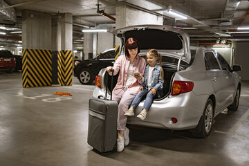 Caucasian mother and little daughter sitting in trunk of modern car in underground parking lot....
