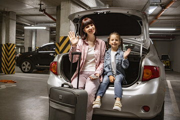 Young woman with child holding traveling suitcase using phone for planning vacation trip. Caucasian mother and little daughter sitting in trunk of modern car in underground parking lot.
