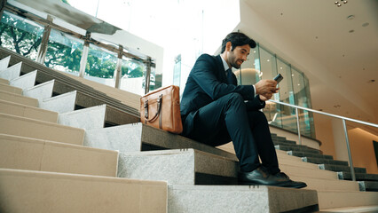 Smart project manager looking at mobile phone while sitting at stairs. Attractive caucasian...