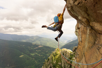 A man is climbing a rock wall with a rope. The man is wearing a yellow jacket and blue jeans....