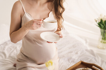  pregnant woman in silk peignoir on a bed among flowers, bouquets of tulips in a vase. Breakfast in bed and coffee, croissant closeup no face. gentle studio pregnancy photo shoot