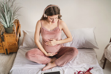 Young attractive pregnant woman listens to music on headphones while sitting on the bed in the bedroom. Concept of early child development, music in the womb. Relaxation and stretching for pregnant wo