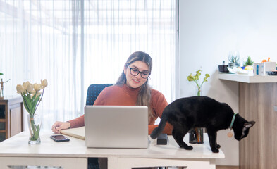 young latin woman working in her home office on her laptop next to a black cat on the desk
