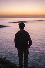 Silhouette of a young man in a burgundy hoodie gazes at the serene Atlantic Ocean and Estelas islands from Monteferro's Rosa de los Vientos viewpoint at sunset, with orange and purple skies.