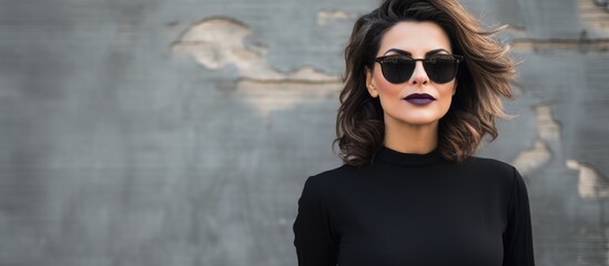 A woman with layered hair wearing sunglasses and a black turtleneck stands in front of a wall, smiling and gesturing with her sleeve
